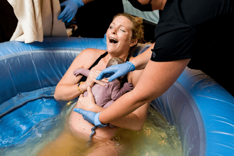 A person has just given birth, and is sitting in a birthing pool, being assisted by a midwife wearing gloves. The person is holding a newborn infant to their chest.