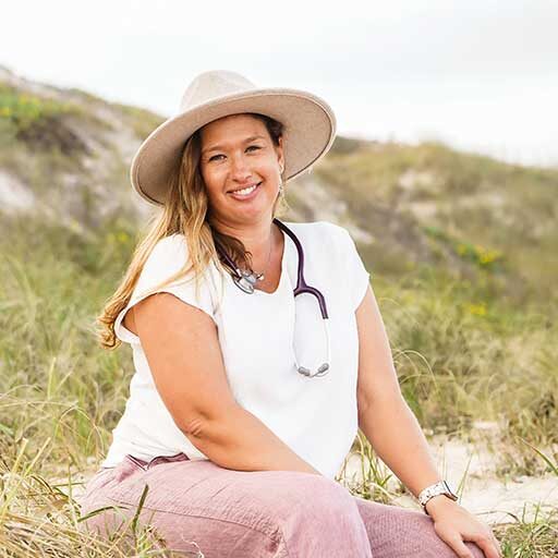 Tiffany Martin Midwife sitting on dune in hat, white shirt and pink pants.